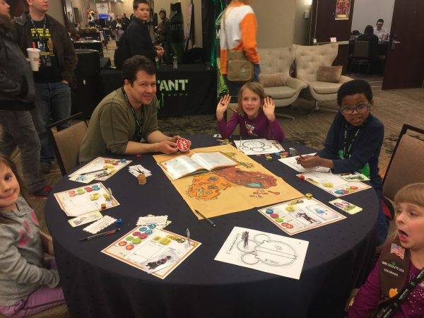 A group of kids sit around a tablew playing a role-playing game. An adult is running the game, and the kids are in mixed states of excitement.