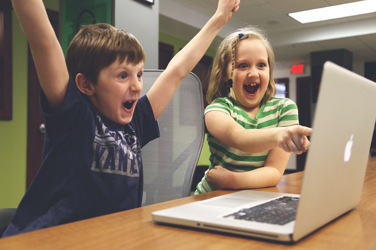 Two children sitting at table in front of a laptop, cheering at an unseen success on the screen