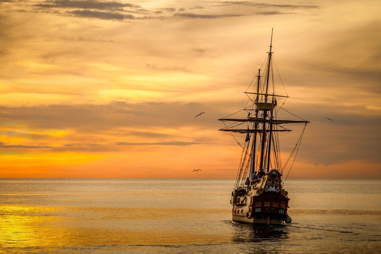A wooden sailing ship heads off on the water towards the sunset.