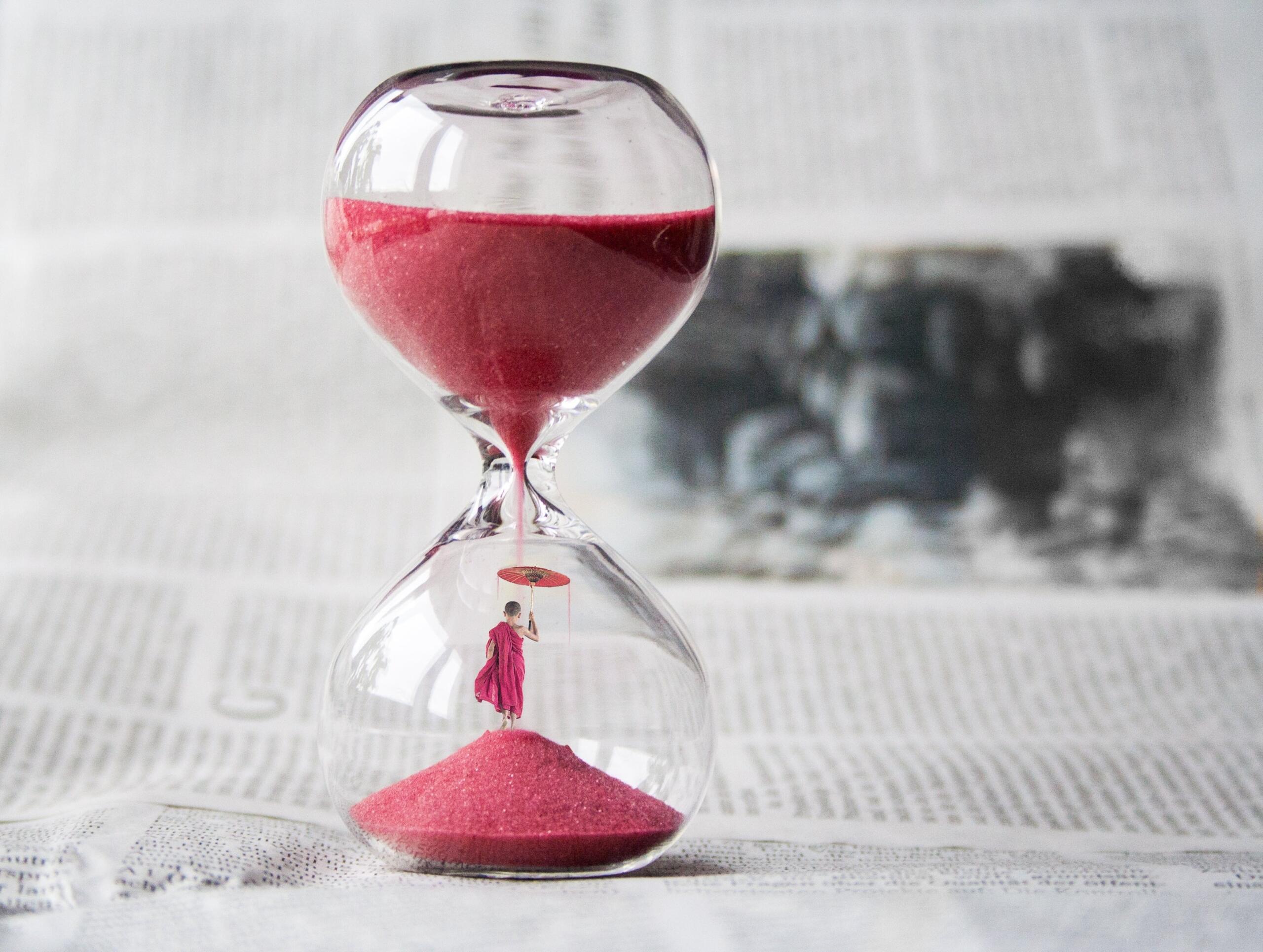 An hourglass with a person inside the bottom of the glass with an umbrella as the grains of sand falling onto the umbrella.
